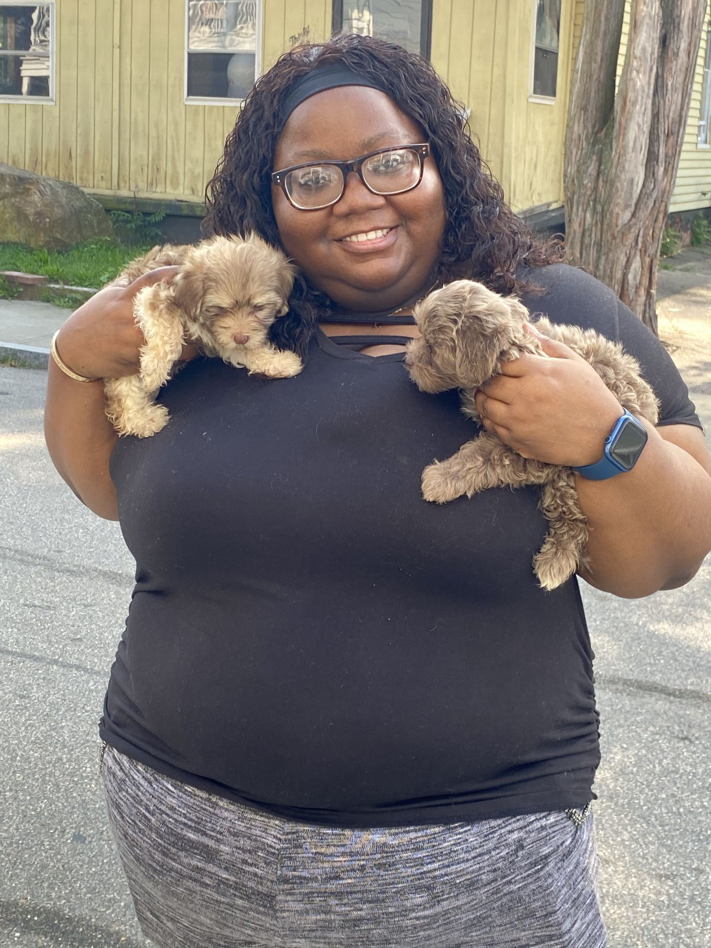 A smiling new owner holding two cream Toy Poodle mix puppies outdoors